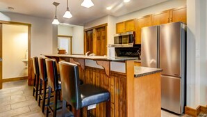 A kitchen with wooden cabinets, a stainless steel refrigerator, a microwave, and a bar area with chairs. - Warm Mountain View Retreat (Canmore)