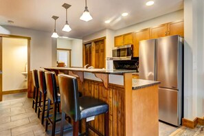 A kitchen with wooden cabinets, a stainless steel refrigerator, a microwave, and a bar area with chairs. - Warm Mountain View Retreat (Canmore)
