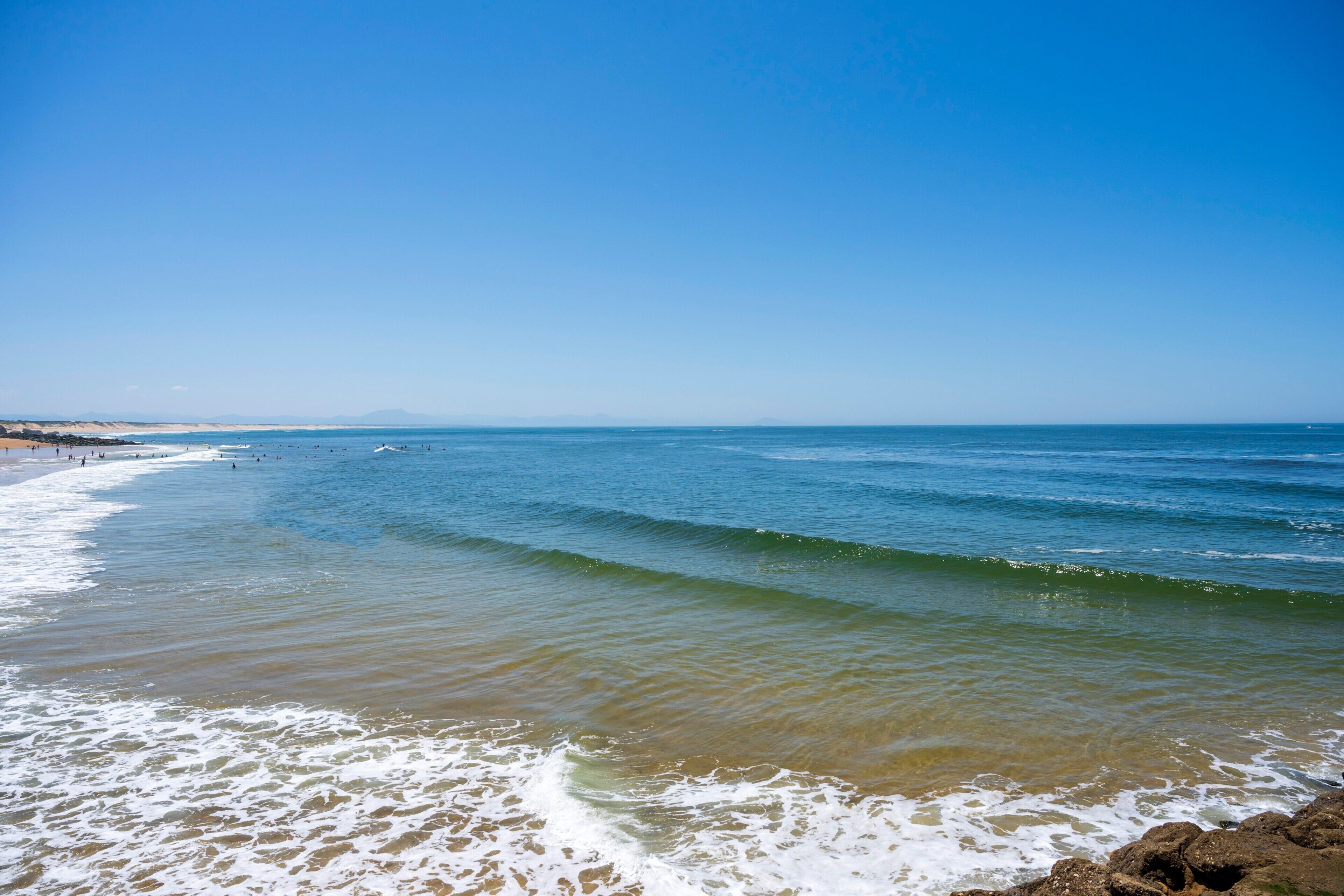 Une plage avec des vagues, un ciel dégagé et des montagnes à l’horizon.