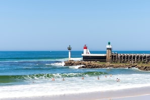 A coastal scene with a lighthouse and pier, waves crashing onto the shore, and people swimming and standing on the rocks.