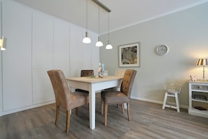 A dining area with a white table, four chairs, pendant lights, a clock, and a small shelf with decorative items. - The Island Seaside Apartment 6 (Timmendorfer Strand)