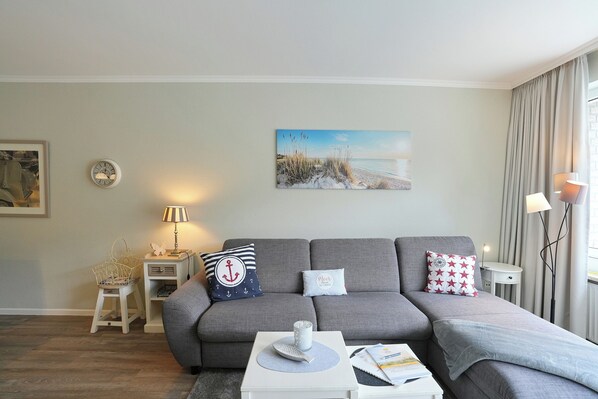 Ein modernes Wohnzimmer mit einem grauen Sofa, einem kleinen Beistelltisch, einer Lampe, einem gerahmten Bild an der Wand und einem Fenster mit Vorhängen.