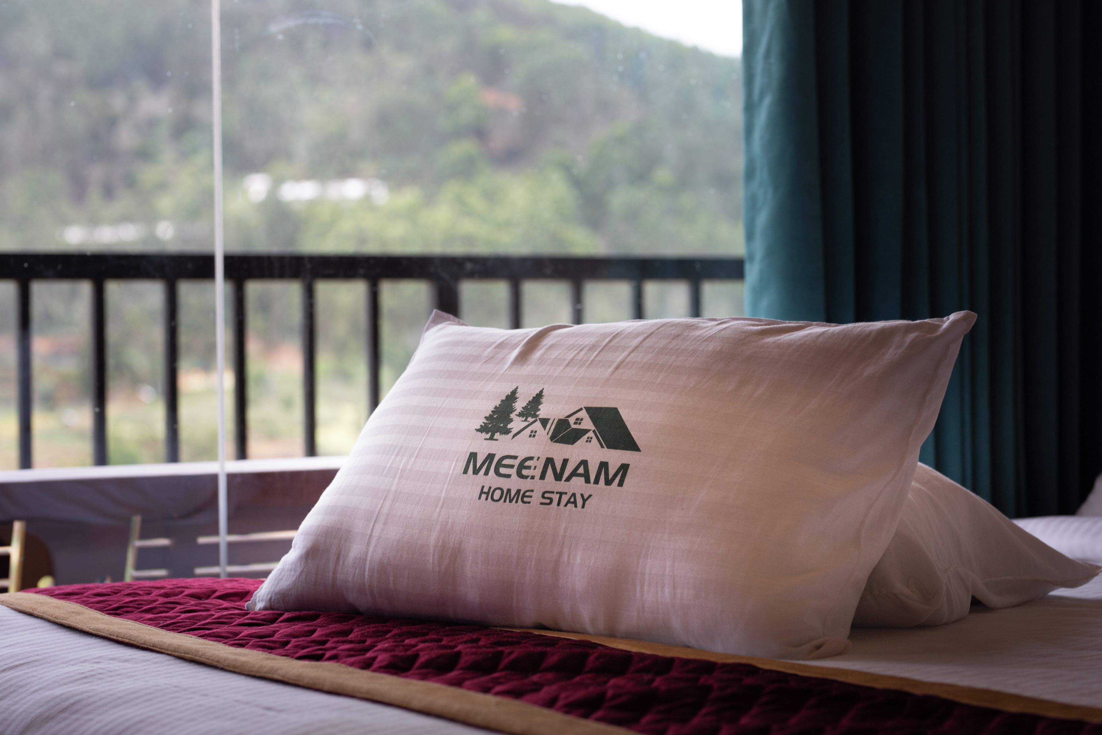 A bed with a pillow featuring a logo and text, a window with a view of greenery, and a curtain.