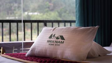 Una cama con almohada que muestra un logotipo y texto, una ventana con vista a follaje, y una cortina.