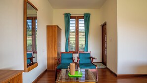 Una habitación con piso de madera, una mesa de vidrio con bandeja verde, dos sillas de madera con cojines turquesa, un armario de madera y una ventana con cortinas turquesa.