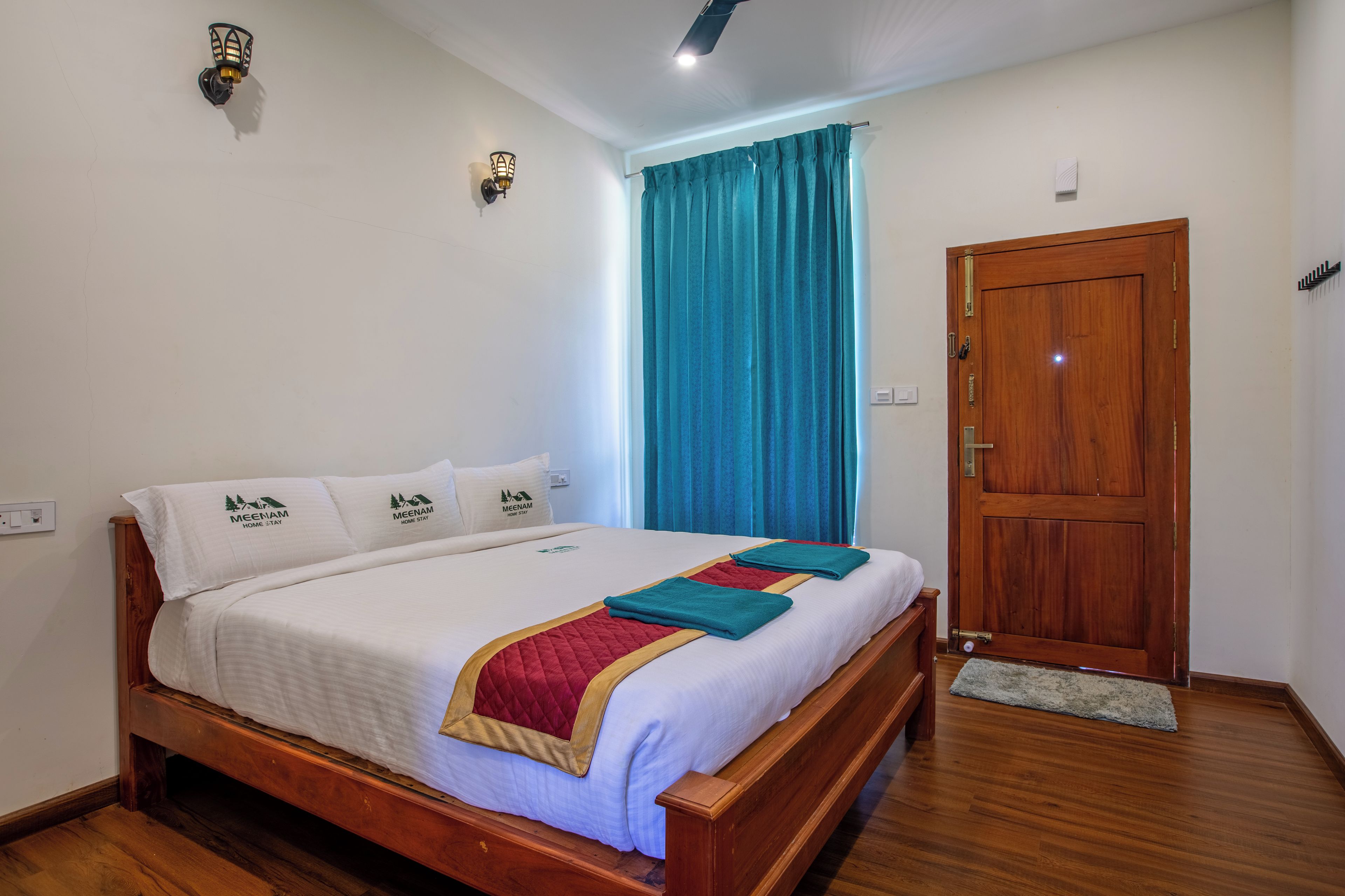 A neatly made bed with a wooden frame, white bedding, and a red and teal blanket. There is a wooden door to the right and a blue curtain partially drawn.