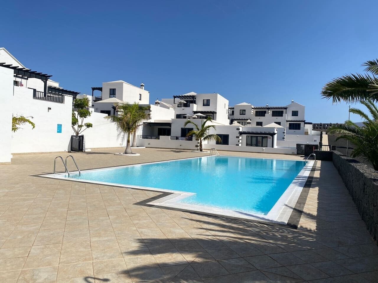 Ein Schwimmbad mit klarem, blauem Wasser, umgeben von einer gefliesten Terrasse und weißen Gebäuden im Hintergrund.