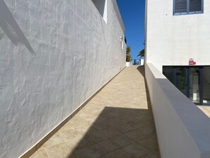 Ein schmaler, von der Sonne durchfluteter Gang mit Fliesenboden, einer weißen Wand auf der einen Seite und einem Gebäudeeingang mit Glastür auf der anderen.