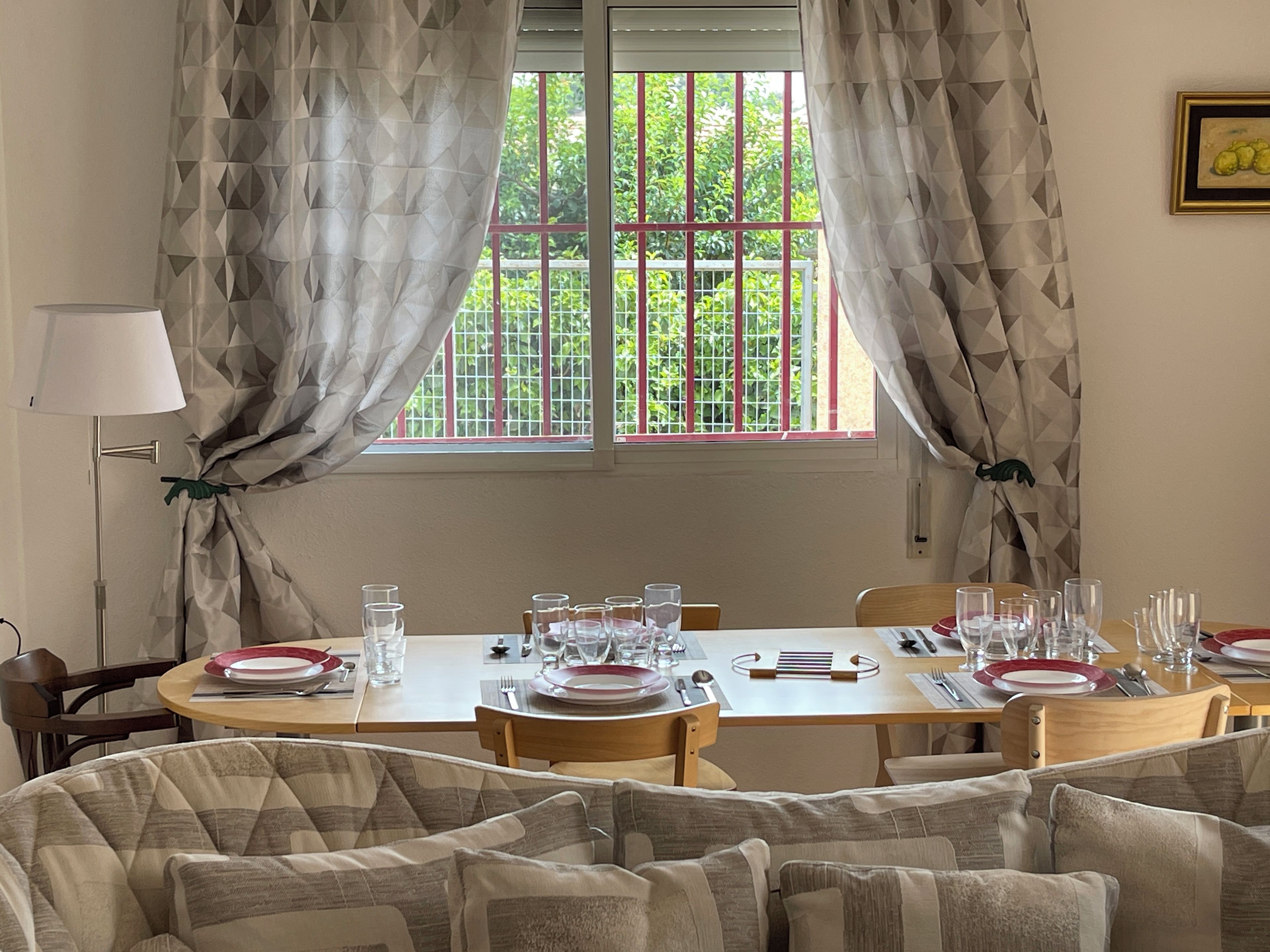 A dining room with a table set for four, curtains, a lamp, and a window with a view of greenery.