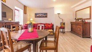 Een eethoek met een houten tafel, stoelen en een rode geruite placemat. Er is een bank met rode kussens, een houten kast en een schilderij aan de muur.