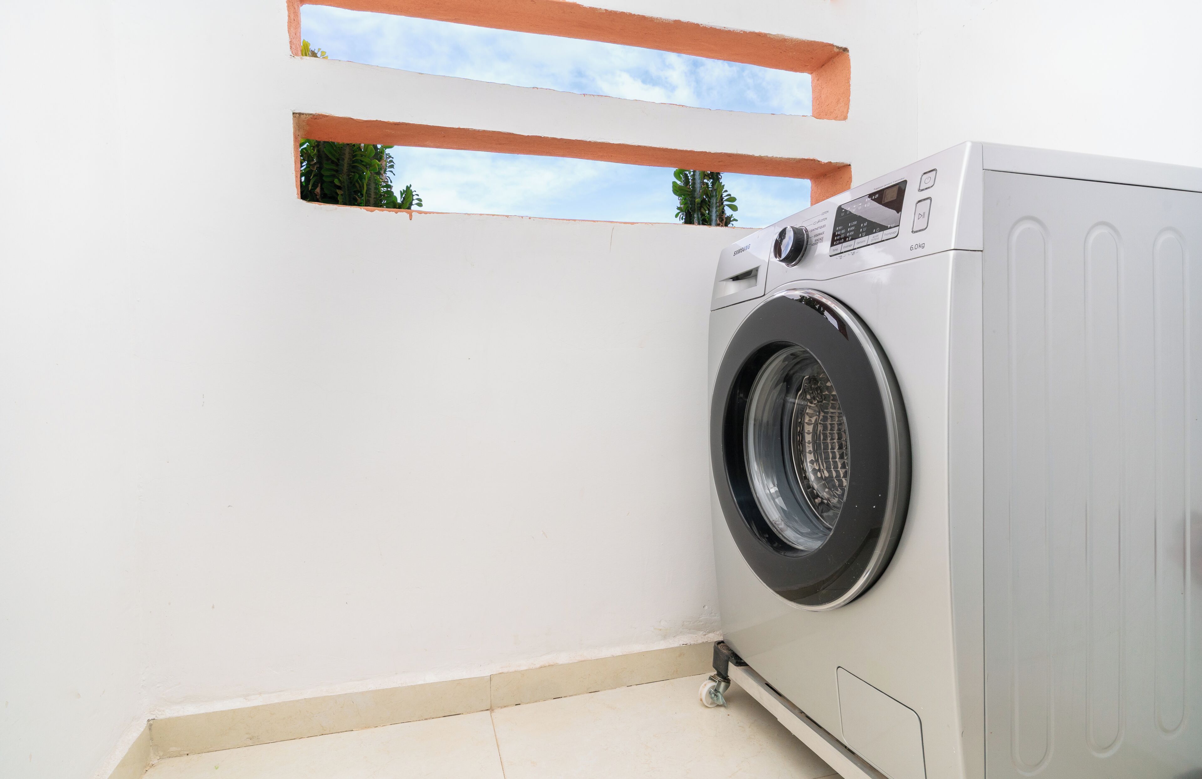Une machine à laver à chargement frontal, placée dans un coin d’une pièce dont le mur est blanc et qui possède une petite fenêtre.