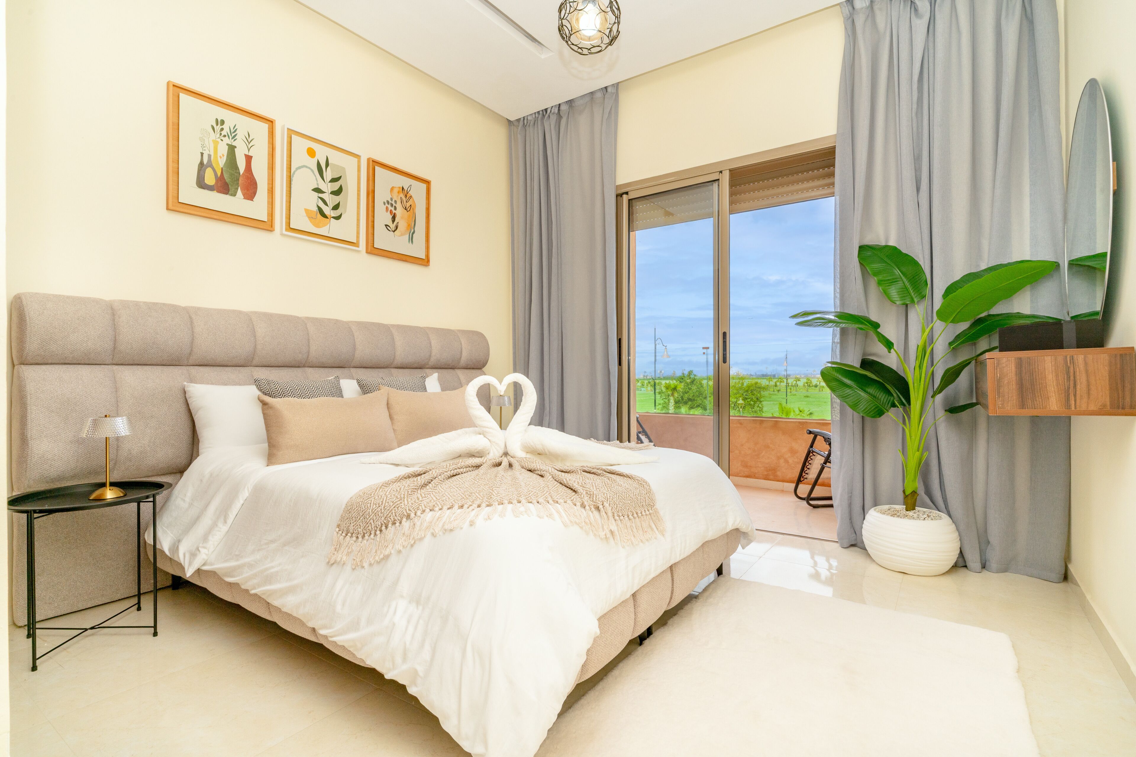 Une chambre à coucher avec un lit, une table de chevet, une lampe, une grande fenêtre avec des rideaux, une plante et des œuvres d’art accrochées au mur.