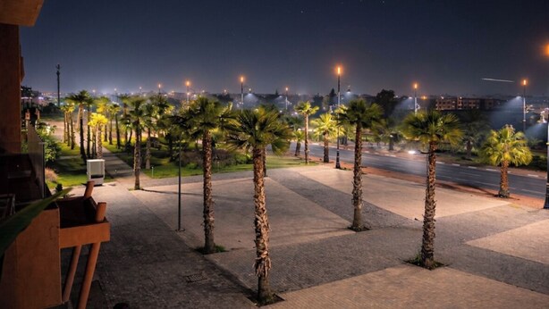 A nighttime view of a paved area with palm trees and streetlights, leading to a road. - Luxury apartment complex with a pool and gym, 10 minutes from Guéliz (marrakech)