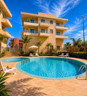 Una piscina con sillas de descanso, cielo azul despejado y un edificio de varios pisos con balcones.