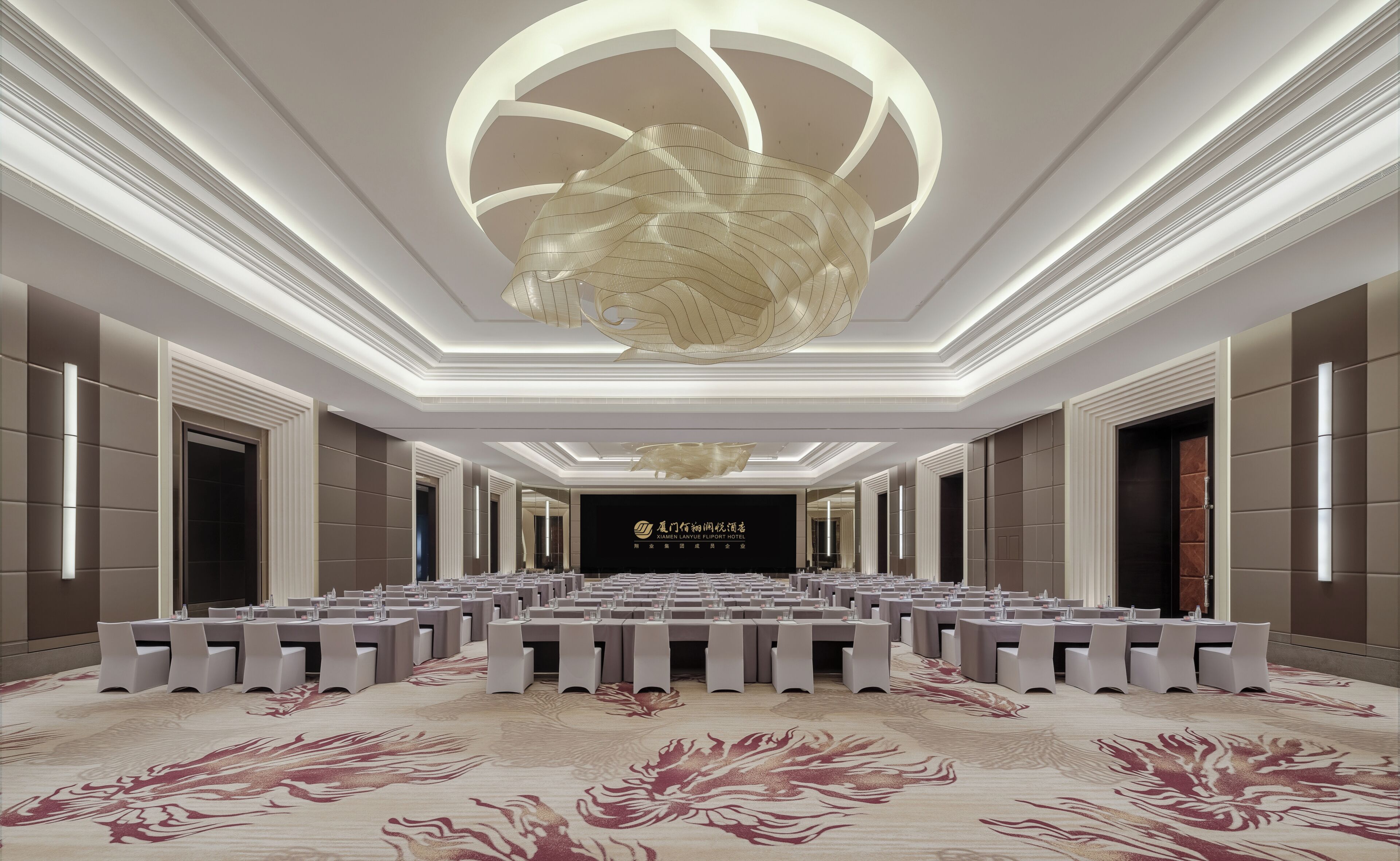 Ein geräumiger Konferenzraum mit einem modernen Kronleuchter, einer großen Leinwand mit Hotelauskunft und einem gemusterten Teppich.