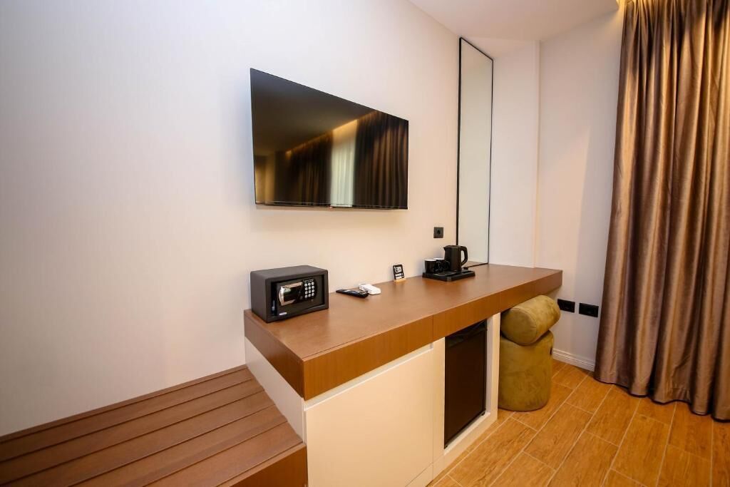 A modern hotel room with a wooden bench, a flat-screen TV mounted on the wall, a small coffee maker, and a trash bin.