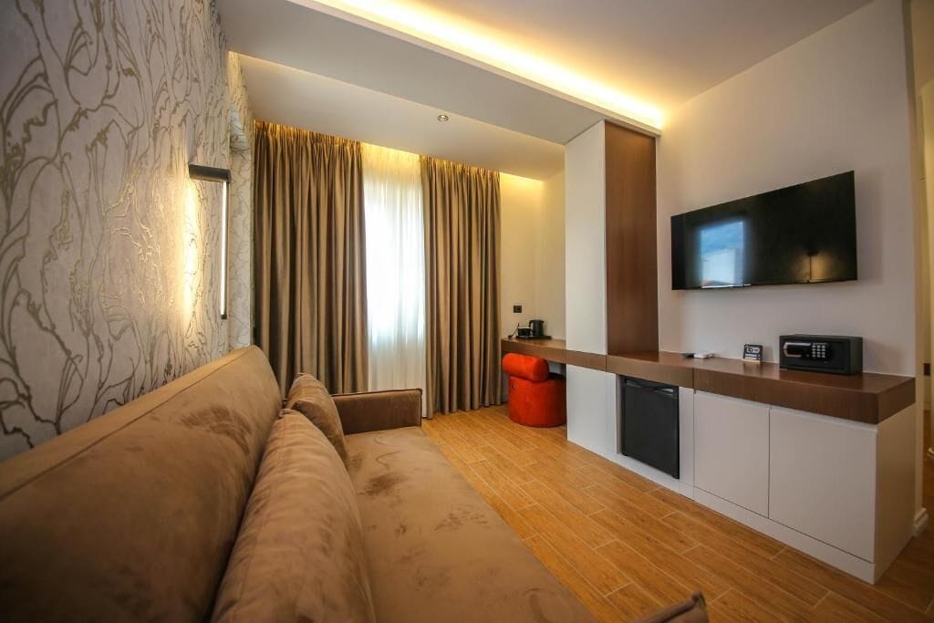A modern hotel room with a sofa, a small kitchen area, a flat-screen TV, and a window with curtains.