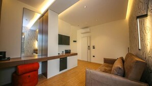 Habitación de hotel moderna con piso de madera, un sofá, un puf rojo, un espejo grande y un televisor de pantalla plana montado en la pared.