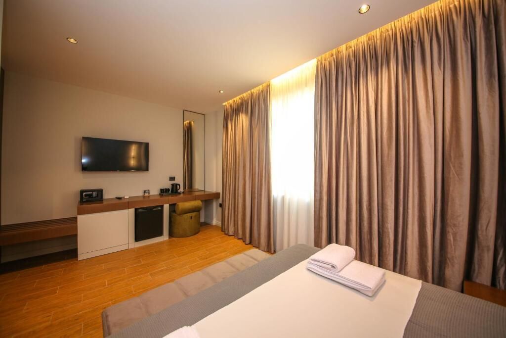 A hotel room with a bed, a desk with a flat-screen TV, a small kitchenette, and a large window with curtains.