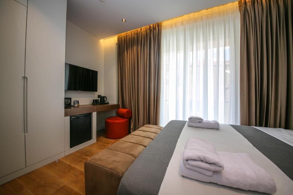 A modern hotel room with a bed, a TV mounted on the wall, a small seating area, and a window with curtains.