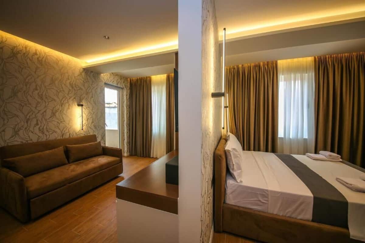 A hotel room with a bed, a sofa, a lamp, and a window with curtains.