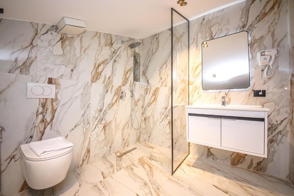 Et badeværelse med en marmorvæg, toilet, brusebad, vaskebord med spejl og en hårtørrer på væggen.