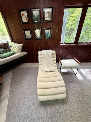 A room with a chaise lounge, a small table, and a bench. There are framed pictures on the wall and a window with a view of trees.