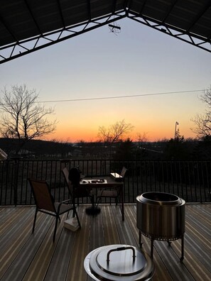 A wooden deck with a table and chairs, a metal grill, and a view of the sunset. - Incredible Spacious Rental with Fantastic Activities for an Unforgettable Family Adventure by the Lake in Cookson, Oklahoma (Cookson)