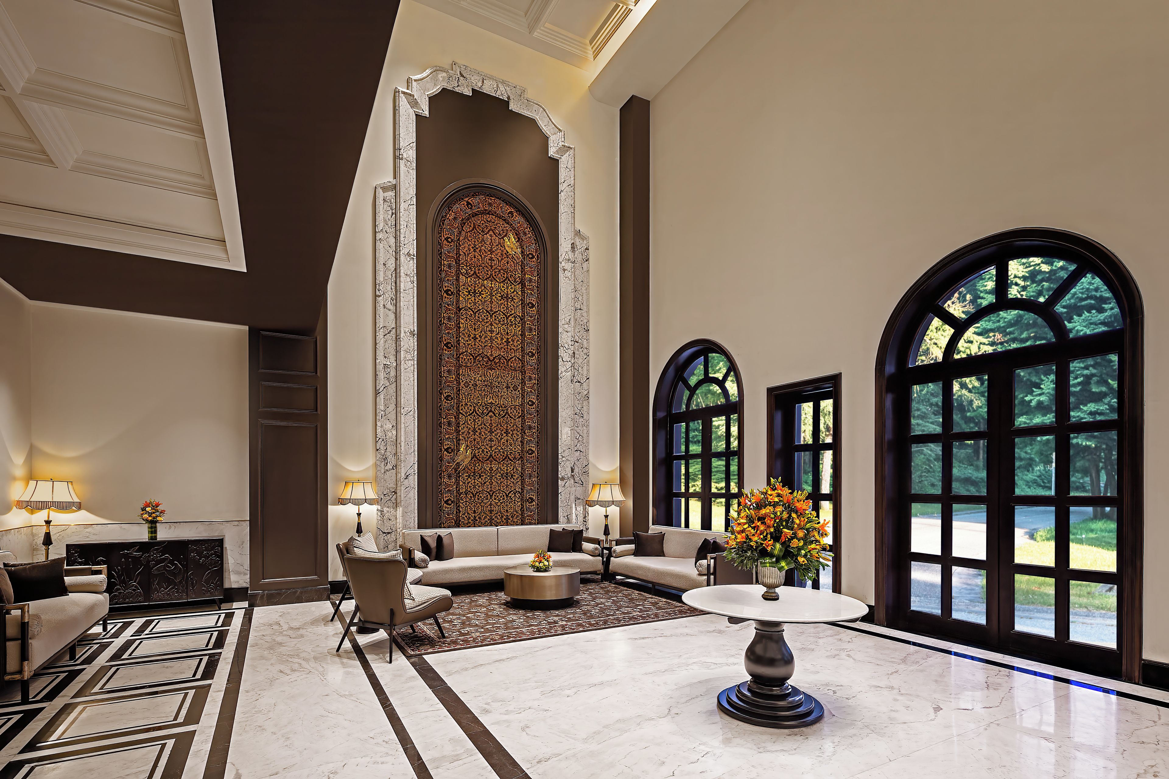 A spacious lobby with a high ceiling, a large ornate wall, a sofa, a coffee table, and a view through large windows.
