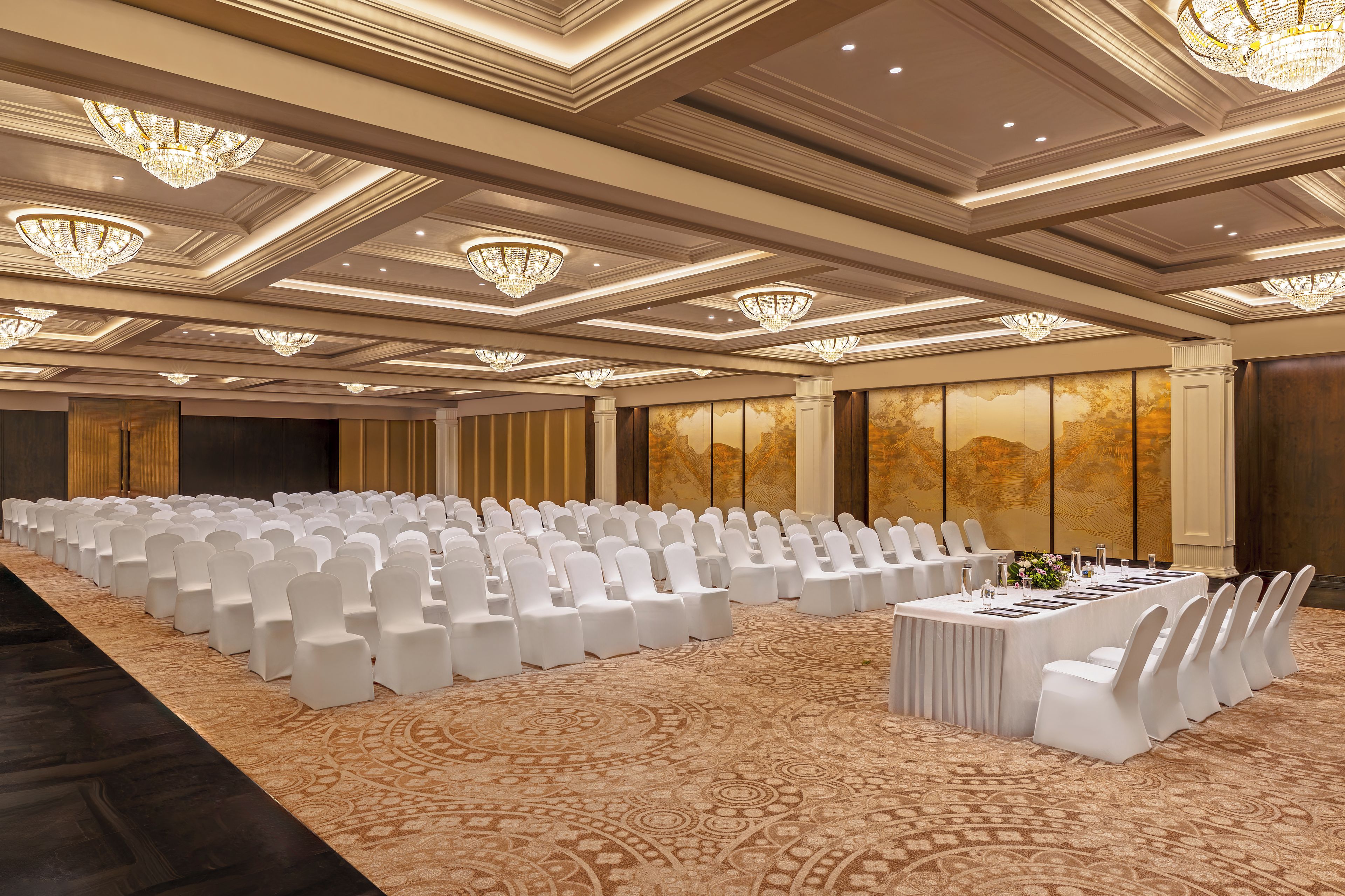 Une salle de conférence avec des rangées de chaises blanches, une longue table agrémentée d’un arrangement floral et des lustres décoratifs.