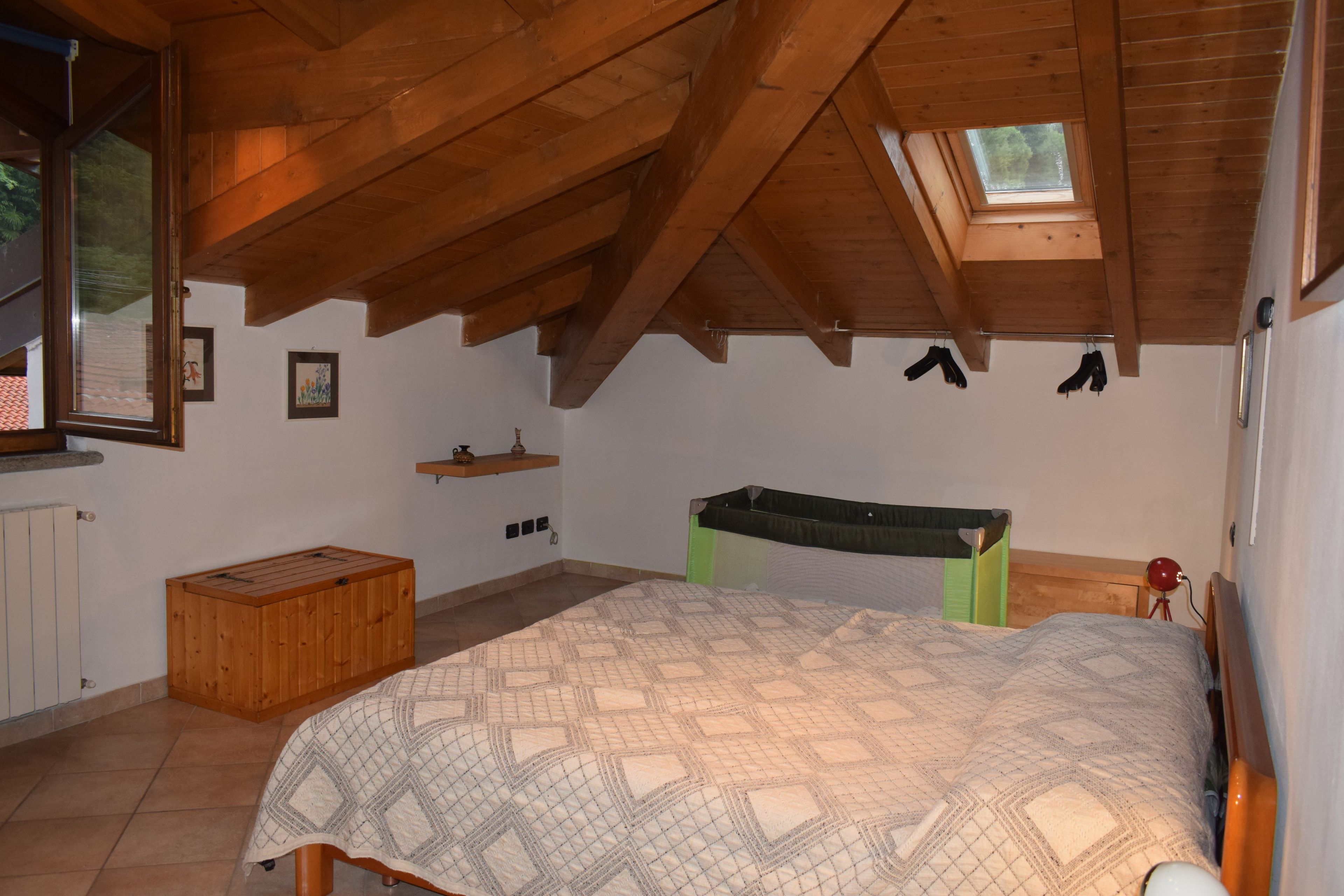Een slaapkamer met een schuine kap, een bed, een houten bankje, een plank met een vaas, en een raam.