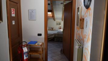 A narrow hallway leading to a bedroom with a bed, a small table with a blue folder, a fire extinguisher, and a radiator.