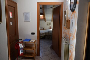 A narrow hallway leading to a bedroom with a bed, a small table with a blue folder, a fire extinguisher, and a radiator.