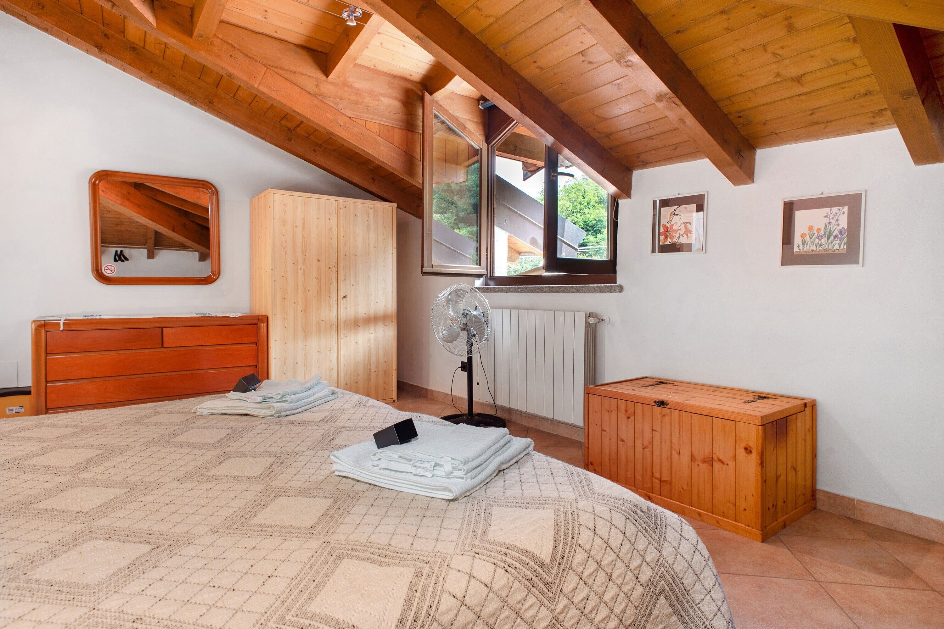 Une chambre avec un plafond en bois, un lit recouvert d’une couvre-lit à motifs, une commode en bois, un miroir et une fenêtre donnant sur un paysage verdoyant.