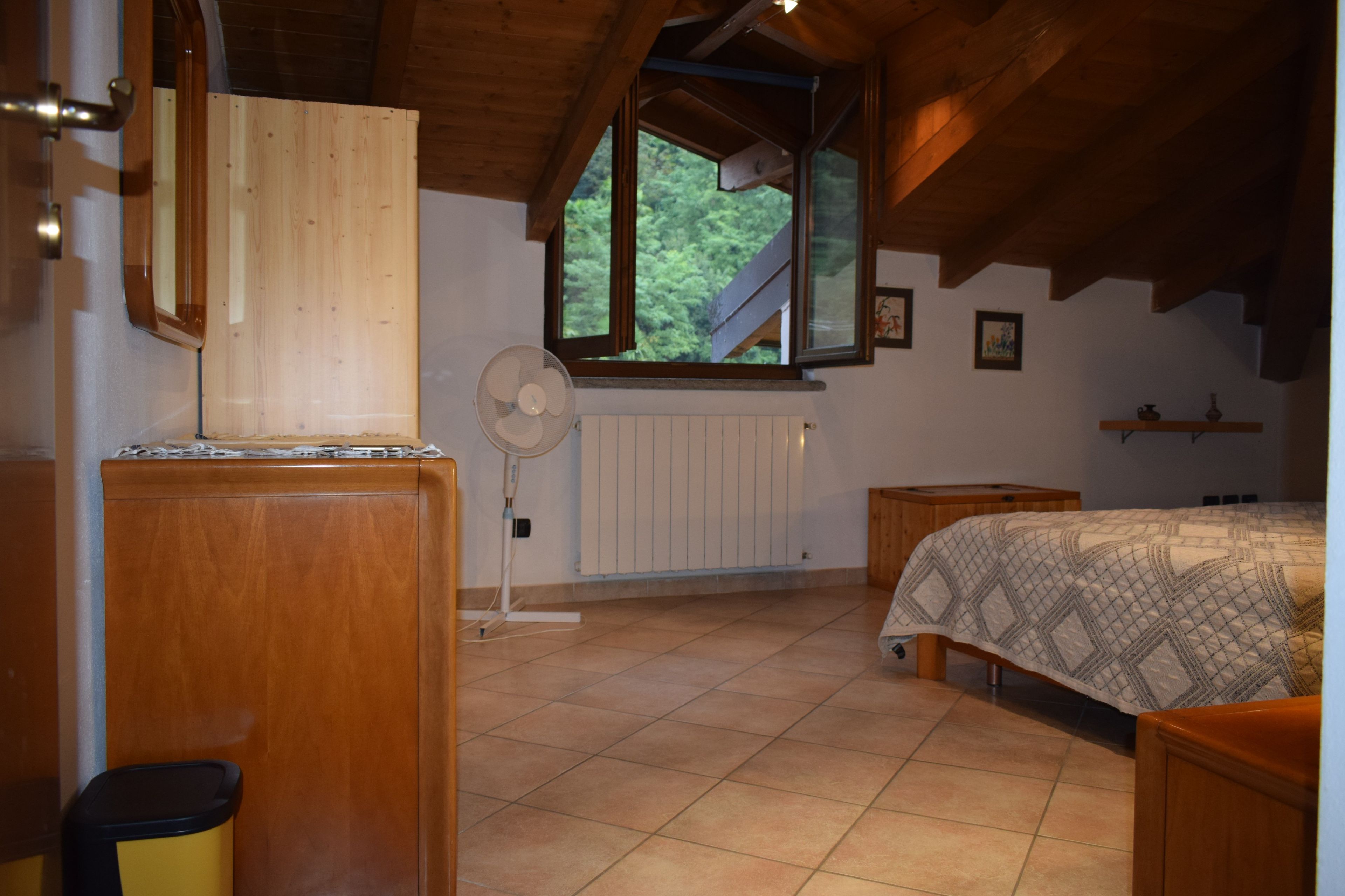 A bedroom with a wooden ceiling, a window, a radiator, a bed with a patterned cover, and a standing fan.