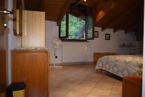 A bedroom with a wooden ceiling, a window, a radiator, a bed with a patterned cover, and a standing fan.