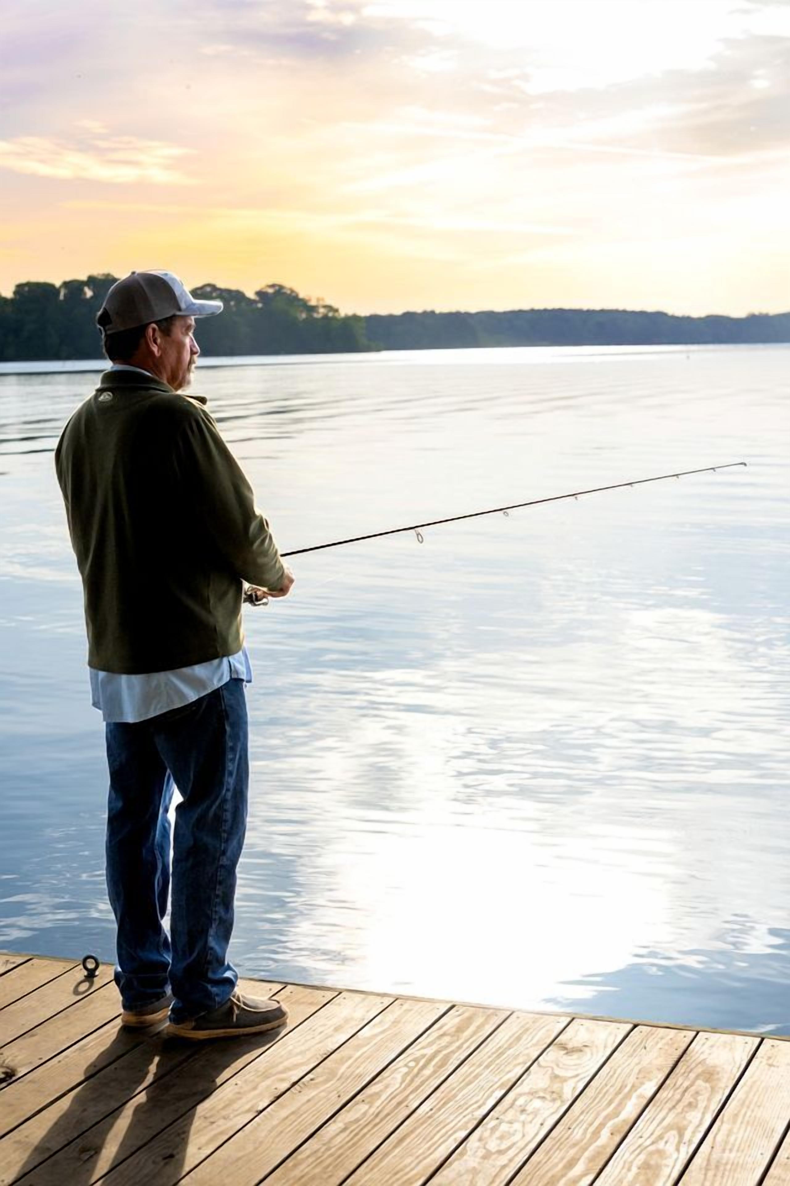 A person standing on a wooden dock holding a fishing rod, facing a calm body of water with a sunset in the background.