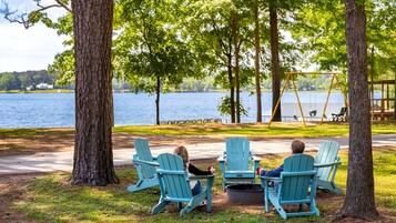 Two people sitting on Adirondack chairs facing a lake, with a swing set and picnic table in the background.