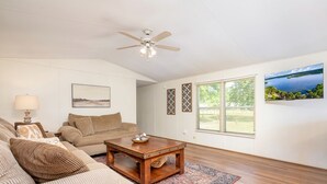 A living room with a ceiling fan, a wooden coffee table, a beige sofa, a patterned rug, and a large window with blinds. - Amazing Vacation Rental in Sabine National Forest on Toledo Bend Lake for a Fantastic Family Escape in Hemphill, Texas (Hemphill)