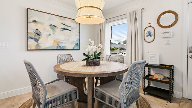 A dining area with a round wooden table, wicker chairs, a hanging light fixture, a painting, and a small shelf with a decorative item. - Private Tropical Oasis • Heated/Cooled Pool (Naples)