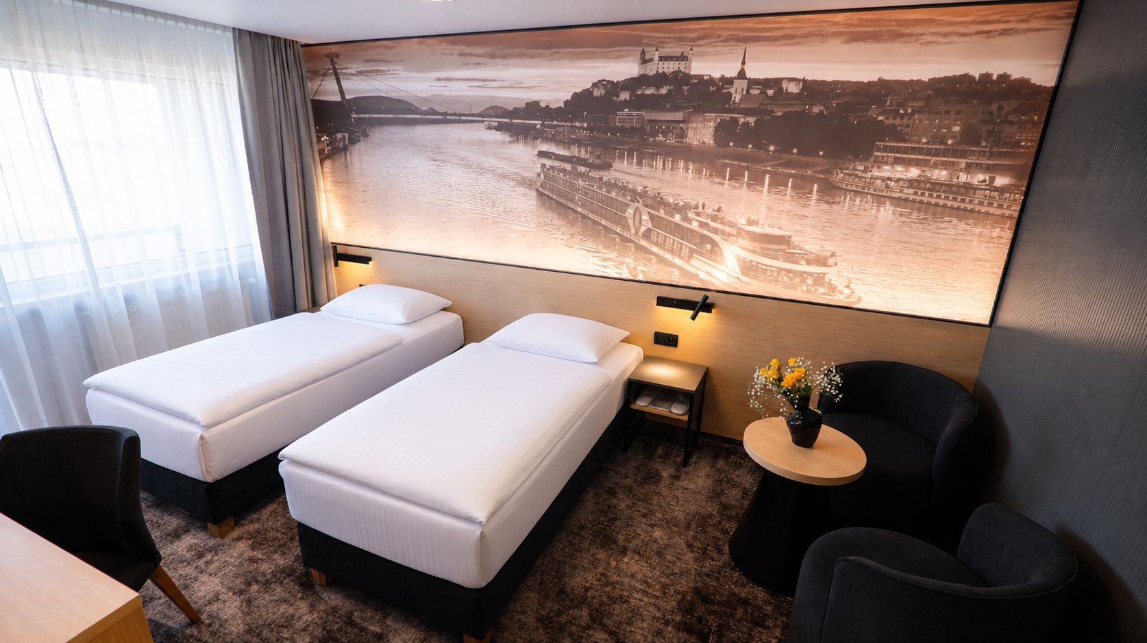 Habitación de hotel con dos camas individuales, una mesita con un florero, una silla y un amplio ventanal con cortinas traslúcidas.