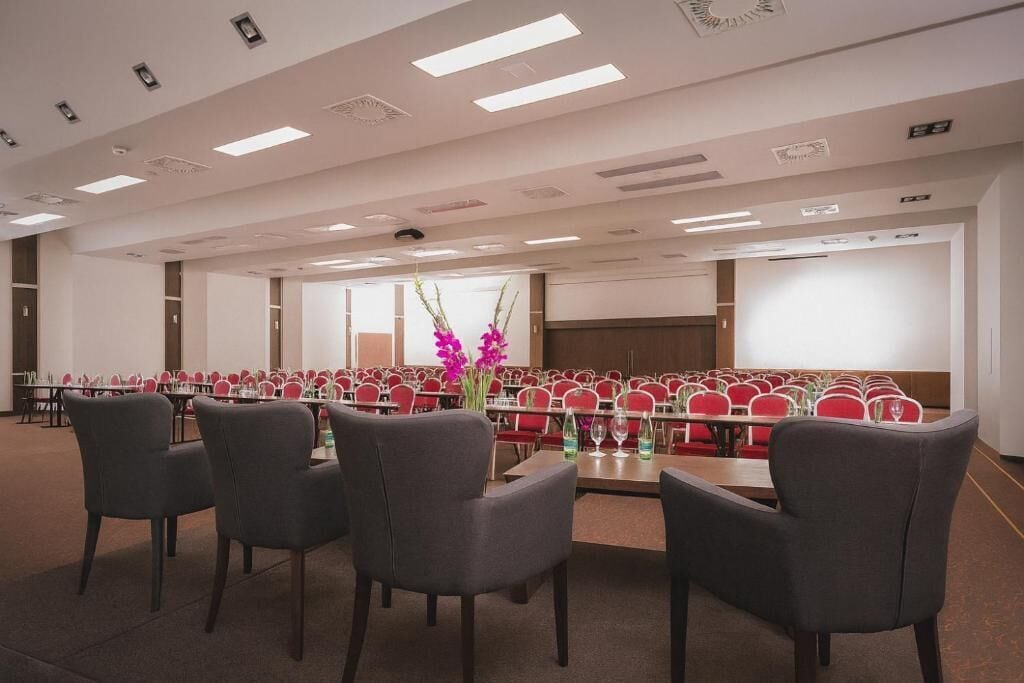 Un salón de conferencias con hileras de sillas rojas, una mesa larga e iluminación empotrada en el techo.