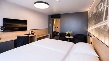 A hotel room with a large bed, a desk with a chair, a small round table with two chairs, and a wall-mounted TV.