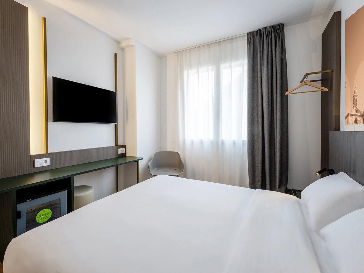 Habitación de hotel con cama, escritorio, silla, televisor y ventana con cortinas.
