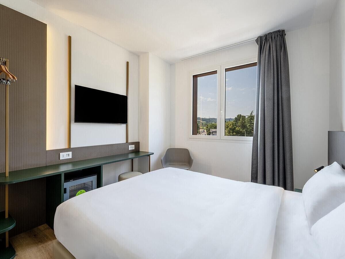 Habitación de hotel con cama, escritorio, silla, televisor y ventana con cortinas.