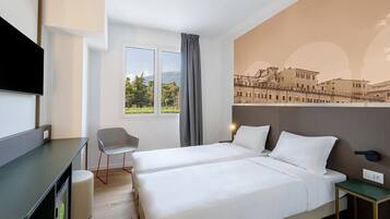 A hotel room with two beds, a desk, a chair, a TV, and a window with a view of buildings.