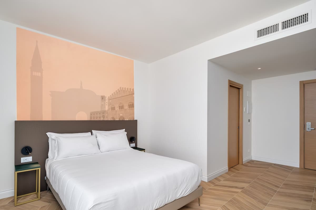 Quarto de hotel com uma cama, mesa de cabeceira, luminária na parede e uma porta.