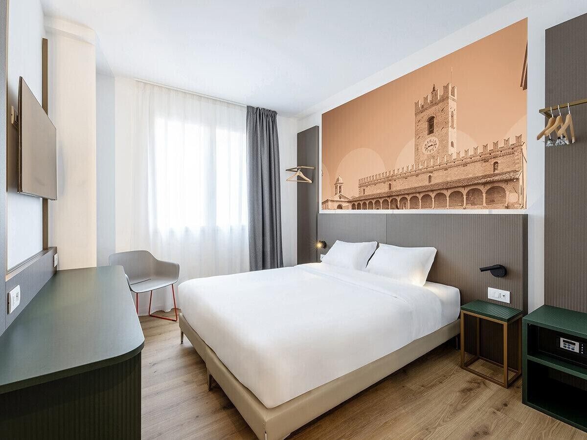 Quarto de hotel com cama, escrivaninha, cadeira, mesa de cabeceira e televisão na parede.