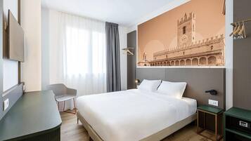 Quarto de hotel com cama, escrivaninha, cadeira, mesa de cabeceira e televisão na parede.