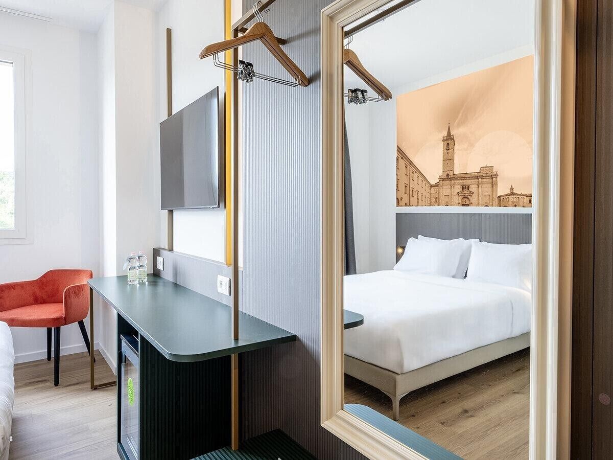 A hotel room with a bed, a desk, a chair, and a large mirror reflecting the room.